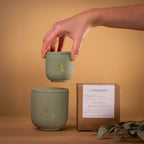Hand holding mini 90g Herbal Sanctuary candle next to large 270g jar and packaging box on wood surface.