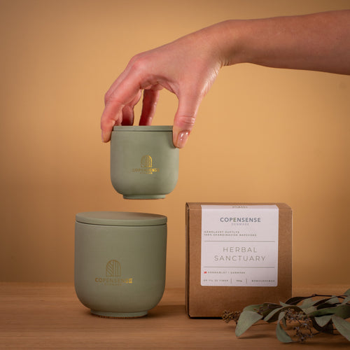 Hand holding mini 90g Herbal Sanctuary candle next to large 270g jar and packaging box on wood surface.